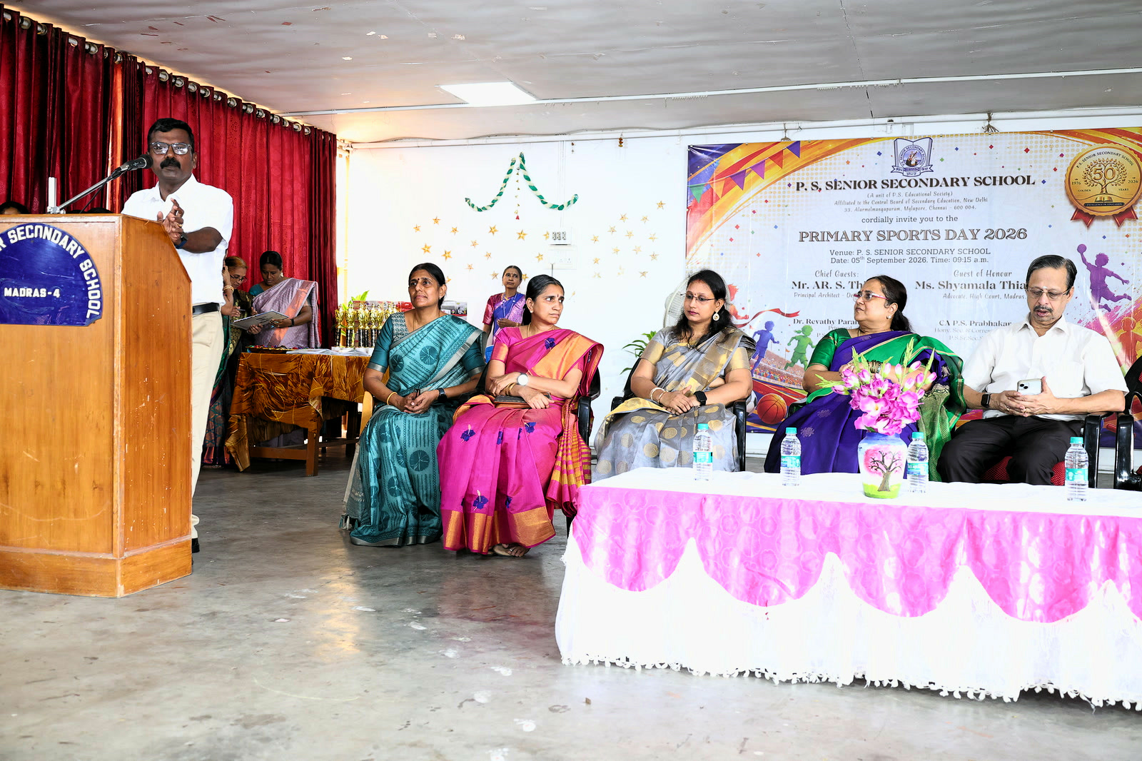 A teacher addressing the audience at the Primary Sports Day 2026 function while guests sit on the dais