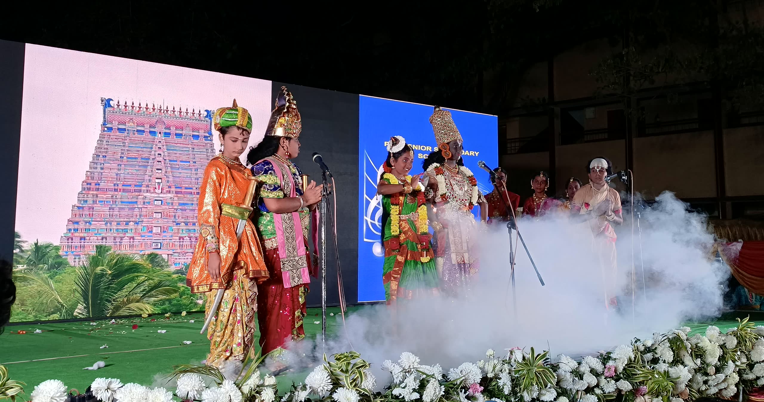 Students in costume performing a scene on the Primary Day stage, with a temple gopuram on the screen behind them