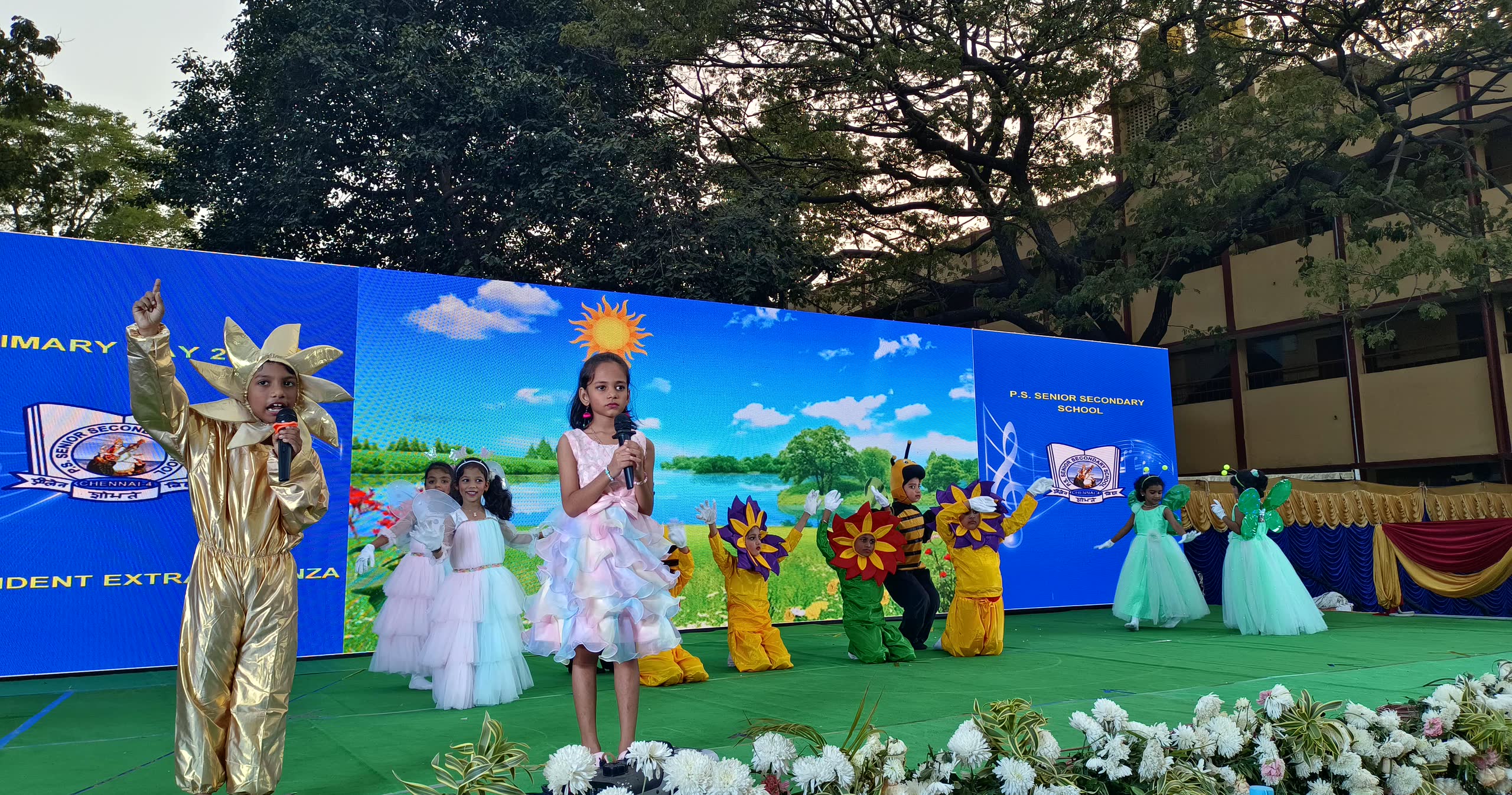 Young children in costume singing on the Primary Day stage in front of a painted lake backdrop